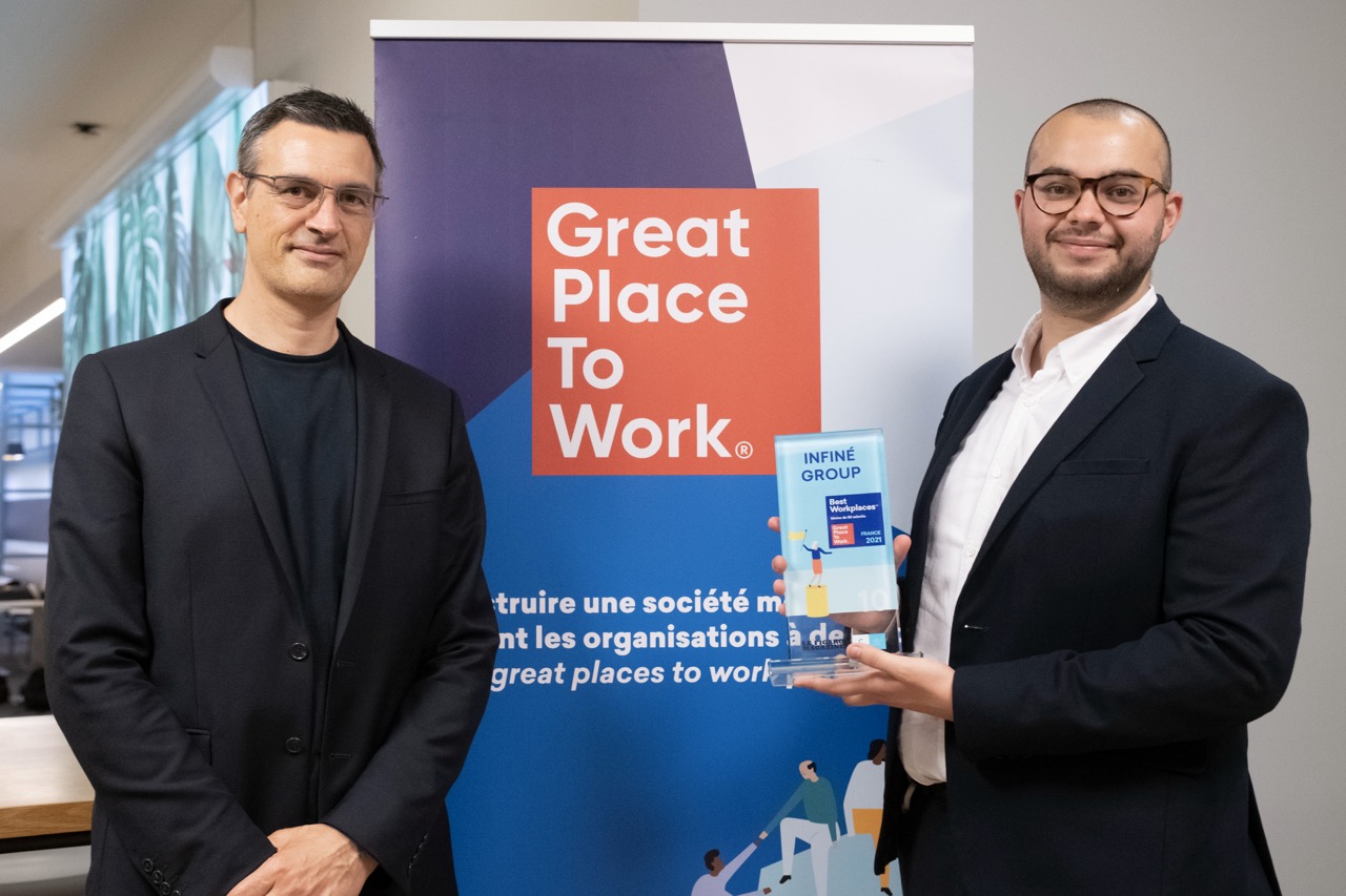 Deux personnes présentent le trophée Best Workplaces 2021 devant le panneau Great Place to Work.